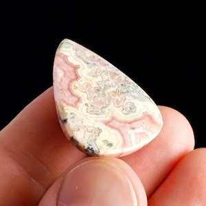 May include: A polished, teardrop-shaped gemstone with a swirling pattern of pink, cream, and gray hues. The stone is held in a hand against a black background, showcasing its natural colors and unique veining.