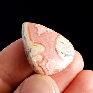 May include: A polished, teardrop-shaped gemstone with a swirling pattern of pink, white, and beige hues. The stone is held in a hand against a black background, showcasing its natural colors and textures. The gemstone's surface is smooth and reflective.