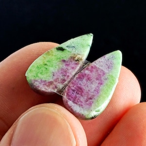 Op de afbeelding: Twee gepolijste edelstenen, elk met een hartvorm, worden getoond. De stenen hebben een levendig kleurenpalet van groen, roze en een vleugje zwart. De edelstenen worden in de hand van een persoon tegen een donkere achtergrond gehouden.