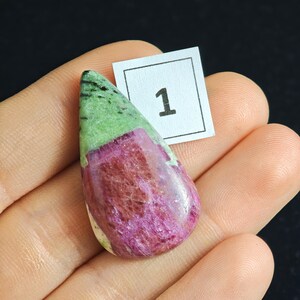 May include: A teardrop-shaped gemstone with a mix of colors. The top portion is green with black specks, transitioning to a vibrant magenta hue. A small white tag with the number "1" is visible.