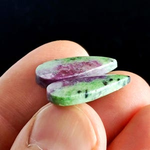 Può includere: Due pietre preziose lucide a forma di goccia. Le pietre presentano un mix di colori, tra cui verde, viola e rosso. Le pietre preziose sono tenute tra le dita su uno sfondo nero. Le pietre hanno una superficie liscia e lucida.