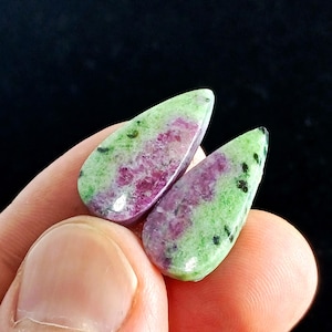 Peut inclure: Deux pierres précieuses en forme de goutte d'eau. Les pierres ont un extérieur vert avec des mouchetures noires et un intérieur rose vif. Les pierres sont tenues entre les doigts sur un fond noir. Ces pierres pourraient être utilisées pour des bijoux.
