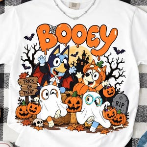 以下が含まれることがあります： BOOEYという文字が入ったカラフルなハロウィンのグラフィックがプリントされた白いTシャツ。吸血鬼や幽霊などの仮装をした4匹の漫画の犬が、カボチャ、RIPと書かれた墓石、木々に囲まれているデザインが特徴です。