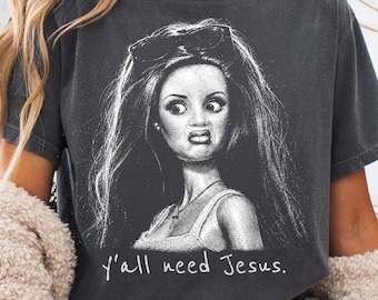 May include: A charcoal grey short-sleeve t-shirt featuring a white graphic illustration of a doll with a sceptical facial expression, wearing sunglasses on her head and a cross necklace. The text y'all need Jesus. is printed in white below.