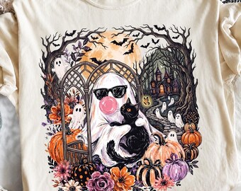 May include: A cream-coloured t-shirt featuring a graphic print of a ghost wearing sunglasses and blowing a pink bubblegum bubble while holding a black cat. The scene includes pumpkins, flowers, a haunted house, bats, and smaller ghosts.