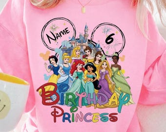 Könnte beinhalten: Rosa Sweatshirt mit farbenfroher Grafik von Disney-Prinzessinnen, einem Schloss und den Worten "Birthday Princess". Das Design enthält die Zahl "6" und Platz für einen Namen. Das Sweatshirt ist leuchtend rosa.