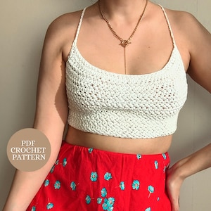 Puede incluir: Un top corto de ganchillo blanco con tirantes finos y un patrón de punto texturizado, combinado con una falda roja con estampado floral azul. Se lleva un collar de cadena dorada con cierre de barra. Una insignia circular muestra el texto PDF CROCHET PATTERN.
