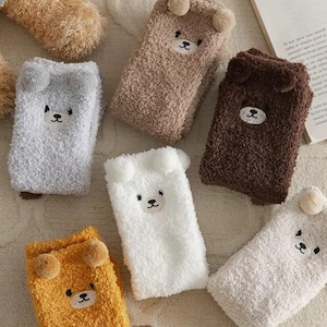 以下が含まれることがあります： グレー、ブラウン、白、黄色など、さまざまな色のふわふわのぬいぐるみソックスのコレクション。各靴下は、刺繍のディテールと小さなポンポンが付いたクマの顔のデザインが特徴で、居心地が良く遊び心のある美学を作り出しています。