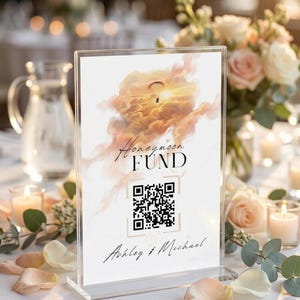 Könnte beinhalten: Transparenter Acrylständer mit der Aufschrift "Honeymoon FUND" in eleganter Schrift, einem QR-Code und den Namen "Ashley & Michael". Das Schild zeigt ein Sonnenuntergangsbild mit einem Gleitschirm. Auf einem Tisch mit Blumengestecken.