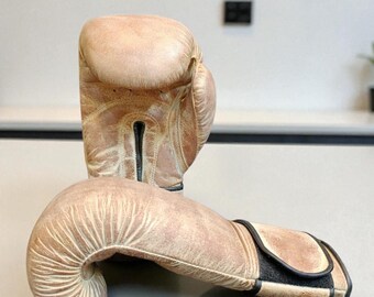 May include: A pair of tan boxing gloves with black accents. The gloves are made of leather and show signs of wear, giving them a vintage look. The gloves are positioned on a light gray surface. The brand logo, a dragon, and the text "HECHO ALMA MADE WITH SOUL" are visible.