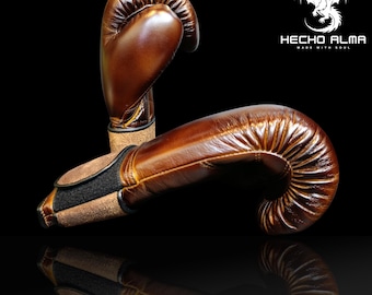 May include: Two brown leather boxing gloves with a textured surface are displayed against a black background. The gloves have a classic design with a padded area for the knuckles and a wrist strap. The logo "HECHO ALMA MADE WITH SOUL" is visible.