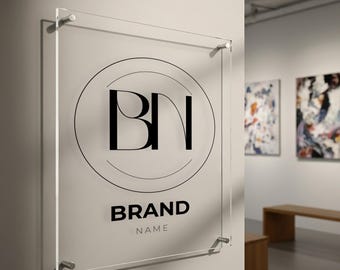 May include: Transparent acrylic sign displaying a black logo with the letters "BN" within a circle. Beneath the logo, the words "BRAND NAME" are printed in a bold, sans-serif font. The sign is fixed to a white wall with silver fixings.