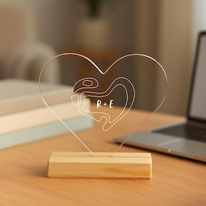 Pode incluir: Uma lâmpada transparente em forma de coração de acrílico com um design de contorno branco e as letras "R + E". A lâmpada está sobre uma base retangular de madeira. Um laptop e livros estão no fundo.