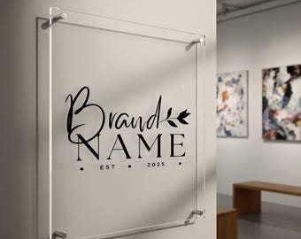 May include: Clear acrylic sign displaying "Brand NAME" in black script, with "EST 2025" below. The sign is mounted on a wall with silver fixings. The sign is in an art gallery.