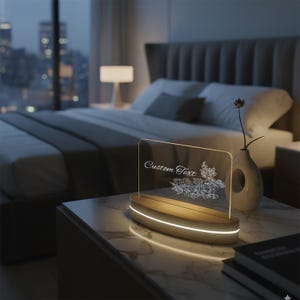 Puede incluir: Letrero acrílico iluminado con las palabras "Custom Text" y un diseño floral. El letrero se encuentra sobre una base de madera con luz. La escena está ambientada en un dormitorio con vistas a la ciudad.