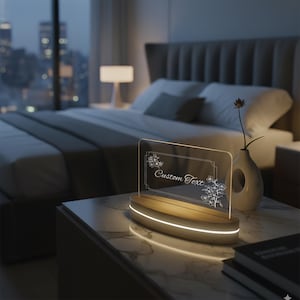 May include: A lighted acrylic sign with the words "Custom Text" and floral designs, set on a wooden base with a glowing edge. The sign is on a marble-topped nightstand in a bedroom with a city view.