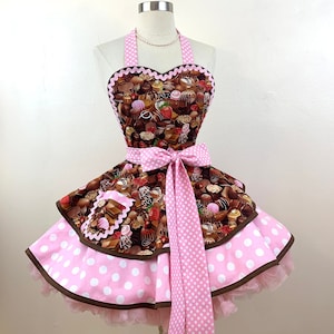 May include: A pink and brown apron with a heart-shaped top featuring a chocolate print. The apron has a pink polka dot bow and a ruffled skirt with a white tulle layer.