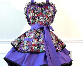 Può includere: Un grembiule a più strati in tessuto nero con una stampa colorata di teschi e fiori sul corpetto e sulla gonna superiore. Include un nastro viola in vita, una gonna inferiore viola, bordi in pizzo nero e una sottogonna nera con volant.