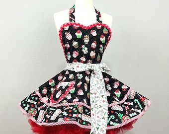May include: A festive black apron with a cupcake pattern, red trim, and a red tulle skirt. The apron features a white ribbon with a botanical design. The apron is displayed on a mannequin.