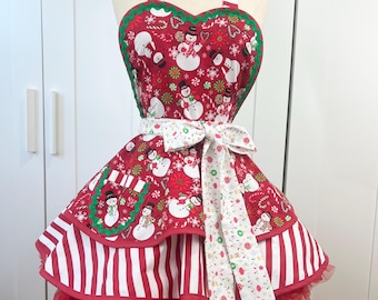 May include: A festive Christmas apron featuring a red background with a snowman pattern, accented with green trim and a white bow. The apron has a striped bodice and a ruffled hem, creating a cheerful holiday look.