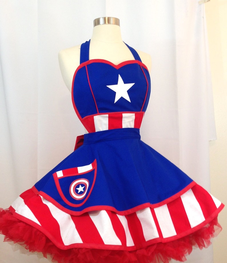Inspired by Captain America,  a red, white, and blue apron with a heart-shaped top and a star in the center. The apron has a pocket on the front with a bullseye design. The lower skirt is a wide red and white stripe. The apron is made of 100% cotton fabric.