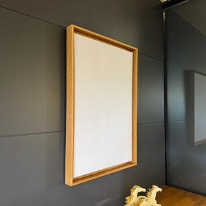 Peut inclure: Une toile rectangulaire vierge avec un cadre en bois naturel. La toile est blanche et le cadre est brun clair. L'œuvre est montée sur un mur gris foncé. L'esthétique générale est minimaliste et moderne.
