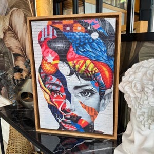 May include: Framed canvas art print depicting a woman's portrait with a vibrant, abstract design. The artwork uses red, blue, orange, and white colours, set against a brick wall background. The frame is made of light brown wood.