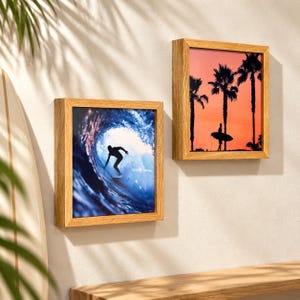May include: Two square wooden frames containing photographic prints. The left frame displays a surfer riding inside a large blue ocean wave. The right frame shows a silhouette of a person holding a surfboard against a vibrant orange sunset with palm trees.