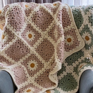 May include: A crochet blanket made of square motifs featuring daisy patterns in dusty rose, sage green, and cream yarn. Each square has a central yellow flower centre surrounded by cream petals and a textured border. The text Karri Krochet Co is visible.