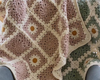 May include: A crochet blanket made of square motifs featuring daisy patterns in dusty rose, sage green, and cream yarn. Each square has a central yellow flower centre surrounded by cream petals and a textured border. The text Karri Krochet Co is visible.