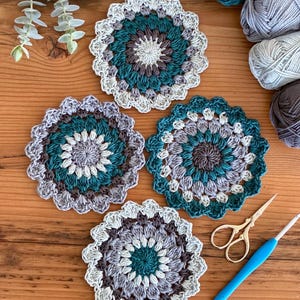May include: Four circular crochet coasters featuring a mandala-inspired pattern in shades of teal, grey, and cream. The coasters have a scalloped edge and are arranged alongside four skeins of yarn, a pair of gold scissors, and a blue crochet hook.