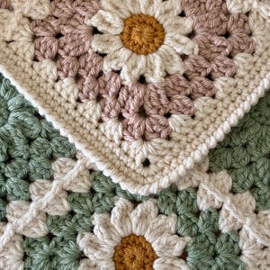 May include: A crochet square featuring a daisy motif with a mustard yellow center, white petals, and a dusty rose background. A second square below features a mustard yellow center, white petals, and a sage green background. Both squares are made of yarn.