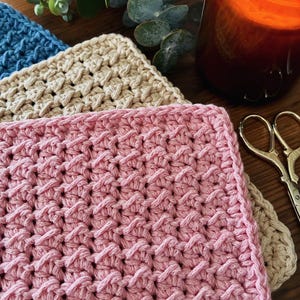 Peut inclure: Un empilement de trois lavettes carrées au crochet en rose, beige et bleu, présentant un motif de points en relief texturé. Les articles sont disposés avec une bougie allumée dans un verre ambré et des ciseaux à broder en métal doré.