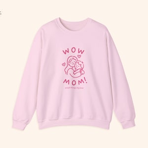 May include: Pale pink sweatshirt featuring the text "WOW MOM!" in pink, with a graphic of a mother and child embracing. The words "small things, big love" are below. The long-sleeved top has a crew neck.