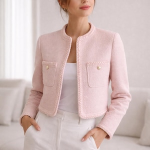 Puede incluir: Chaqueta corta de tweed rosa claro con cuello redondo y dos bolsillos delanteros. La chaqueta tiene botones dorados y ribetes trenzados. La modelo lleva un top blanco y pantalones blancos.