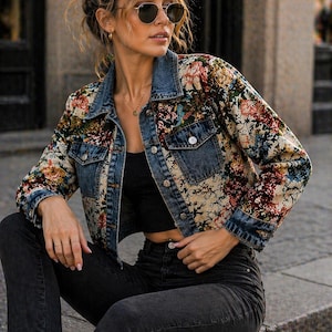 May include: A cropped denim jacket featuring a multi-colored floral tapestry pattern on the sleeves and back panels, contrasted with solid blue denim chest pockets, collar, and front placket, finished with silver-toned metal buttons and cuffs.