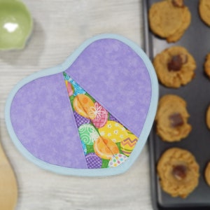May include: A heart-shaped purple and blue pot holder with a colorful Easter egg pattern. The pot holder is made of fabric and has a light blue trim.