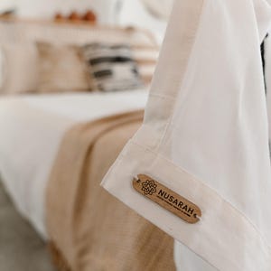 May include: Close-up of a white garment with a wooden tag that reads "Nusarah". The tag has a floral design. The garment is hanging in front of a bed with white linens and a tan blanket.