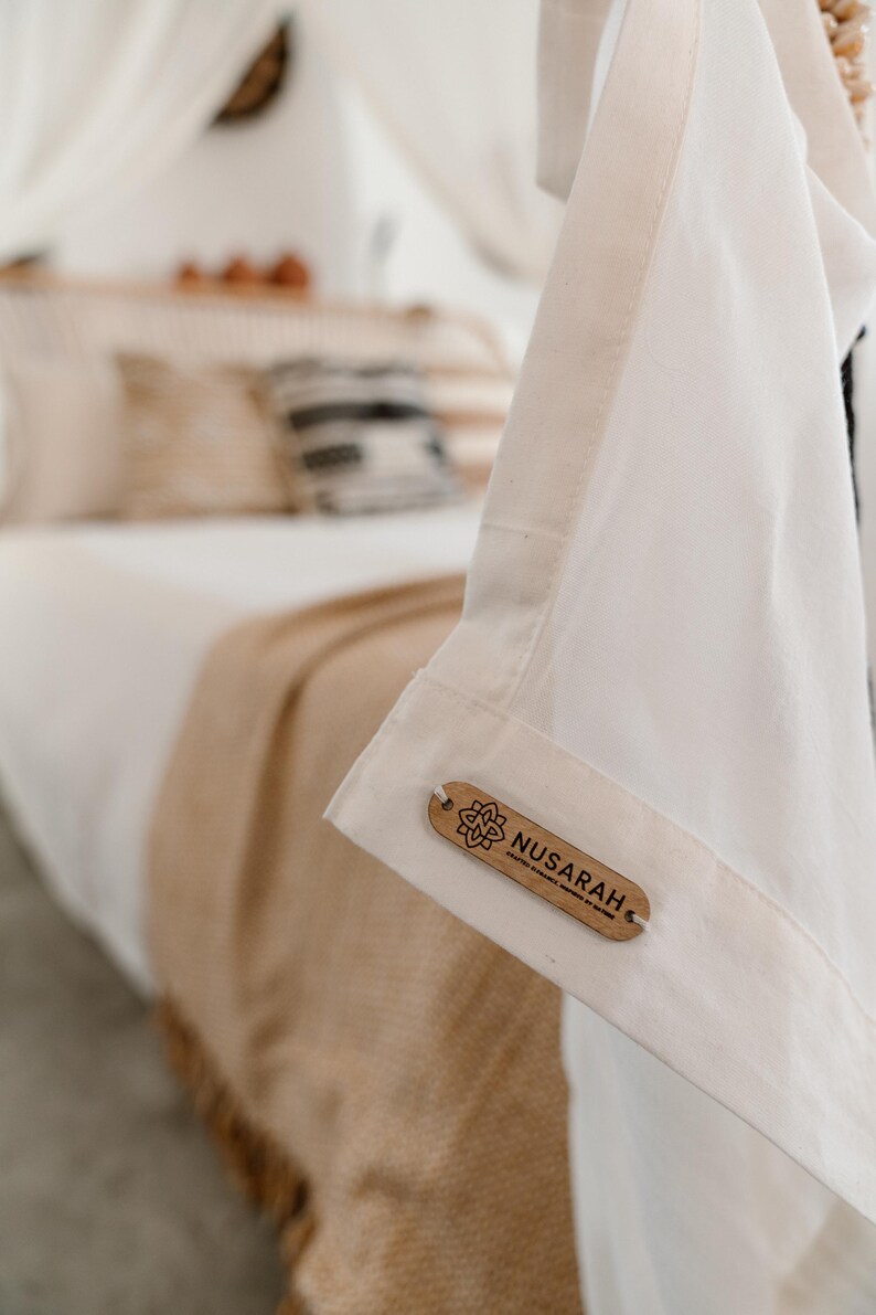 May include: Close-up of a white garment with a wooden tag that reads "Nusarah". The tag has a floral design. The garment is hanging in front of a bed with white linens and a tan blanket.