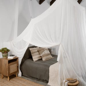 Puede incluir: Una habitación con una cama con dosel blanco. La cama está cubierta con un edredón verde oscuro y almohadas decorativas. Una mesita de noche de madera con frente tejido y una pequeña planta se encuentra junto a la cama. Una alfombra y un taburete tejidos completan la escena.