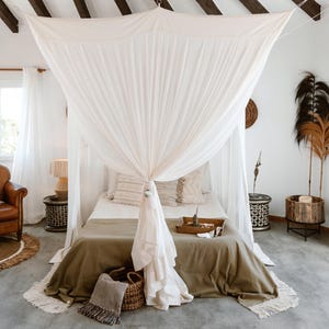 Puede incluir: Un dormitorio con una cama con dosel cubierta con tela blanca. La cama está cubierta con una manta verde y almohadas decorativas. Una cesta tejida y una manta con flecos están al pie de la cama. La habitación tiene una paleta de colores neutros.