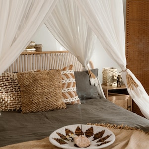 May include: A bedroom scene featuring a bed with a canopy of white sheer fabric. The bed is adorned with decorative pillows in shades of brown and gray. A decorative bowl with a leaf design sits on the bed, alongside a woven throw.