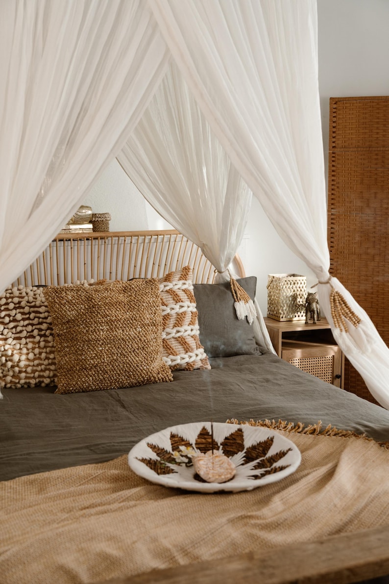 May include: A bedroom scene featuring a bed with a canopy of white sheer fabric. The bed is adorned with decorative pillows in shades of brown and gray. A decorative bowl with a leaf design sits on the bed, alongside a woven throw.