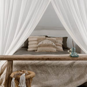 May include: A bedroom scene featuring a bed draped with a neutral-toned linen blanket and pillows. White sheer curtains frame the bed, creating a soft, airy atmosphere. A small woven stool with tassels sits in the foreground, complementing the natural aesthetic.