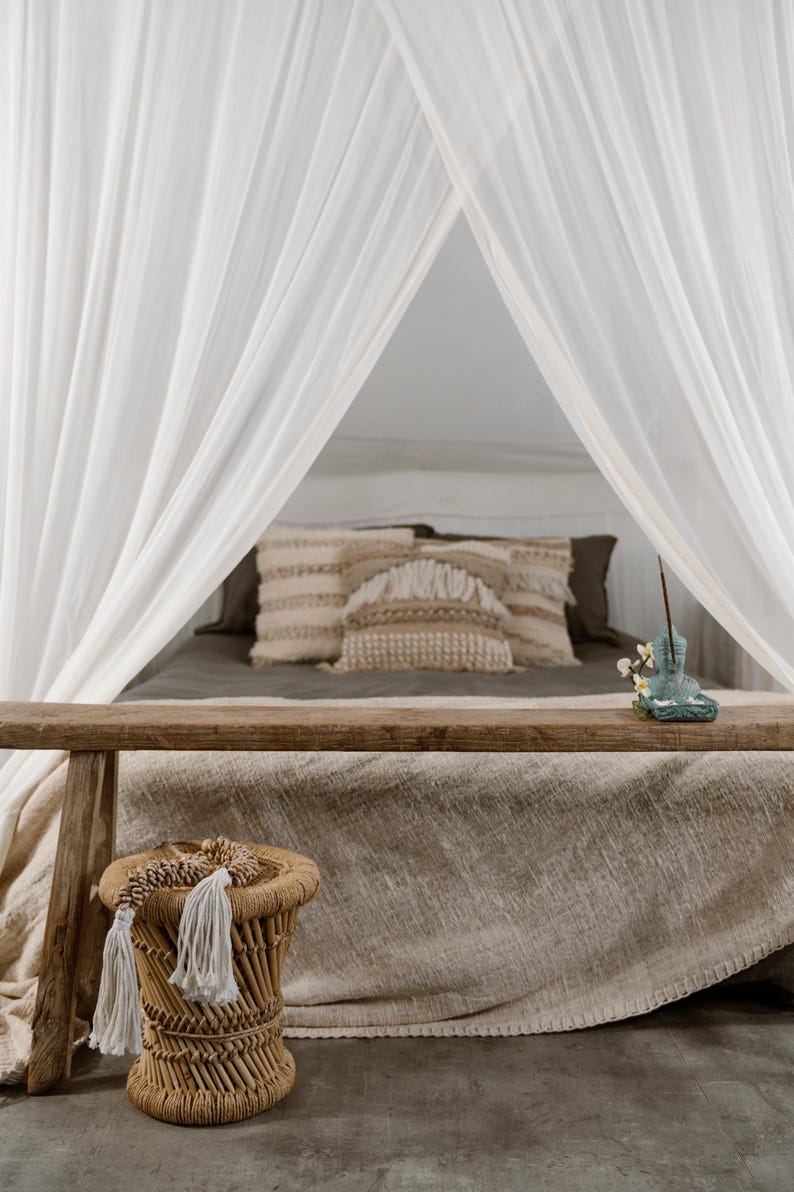 May include: A bedroom scene featuring a bed draped with a neutral-toned linen blanket and pillows. White sheer curtains frame the bed, creating a soft, airy atmosphere. A small woven stool with tassels sits in the foreground, complementing the natural aesthetic.