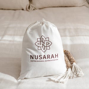 May include: A white drawstring bag with the brand name "NUSARAH" and a floral logo in brown. The bag is on a bed with beige linens, accompanied by a decorative tassel.