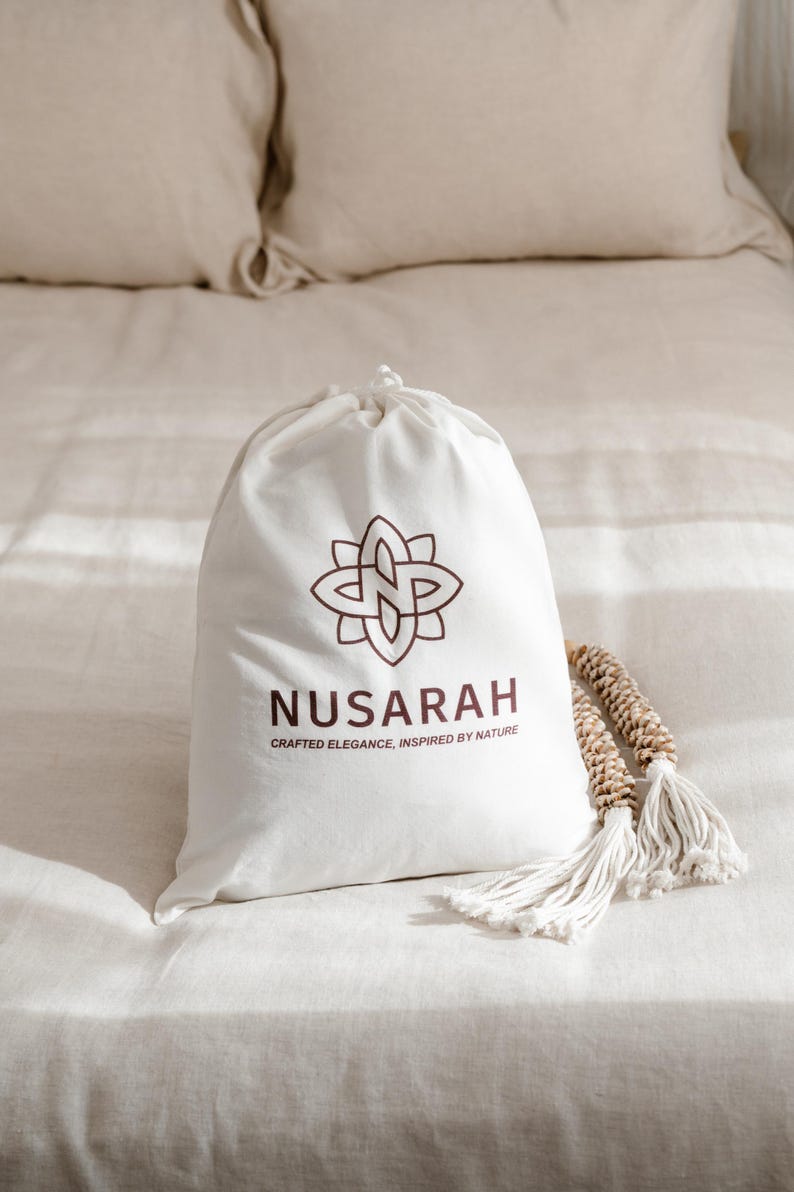 May include: A white drawstring bag with the brand name "NUSARAH" and a floral logo in brown. The bag is on a bed with beige linens, accompanied by a decorative tassel.