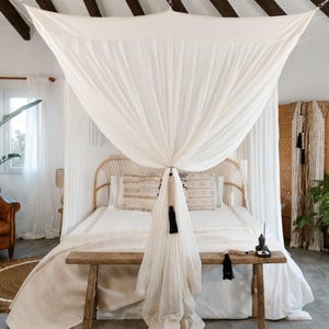 Puede incluir: Un dormitorio con una cama con dosel. La cama tiene un dosel blanco drapeado desde el techo. La cama tiene un cabecero de ratán, ropa de cama blanca y almohadas decorativas. Un banco de madera se encuentra al pie de la cama.