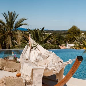 Puede incluir: Una hamaca blanca con estructura de madera está instalada cerca de una piscina. Mantas y cojines beige están sobre la hamaca. Una bandeja con una botella y vasos está cerca sobre una estera tejida. Palmeras y cielo azul en el fondo.