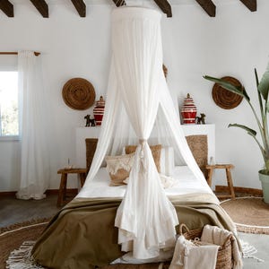 May include: A bedroom with a canopy bed, featuring a white sheer canopy and a bed covered with a green blanket. Decorative items include woven wall hangings, red and white vases, and a large potted plant. A woven rug and basket add to the room's decor.